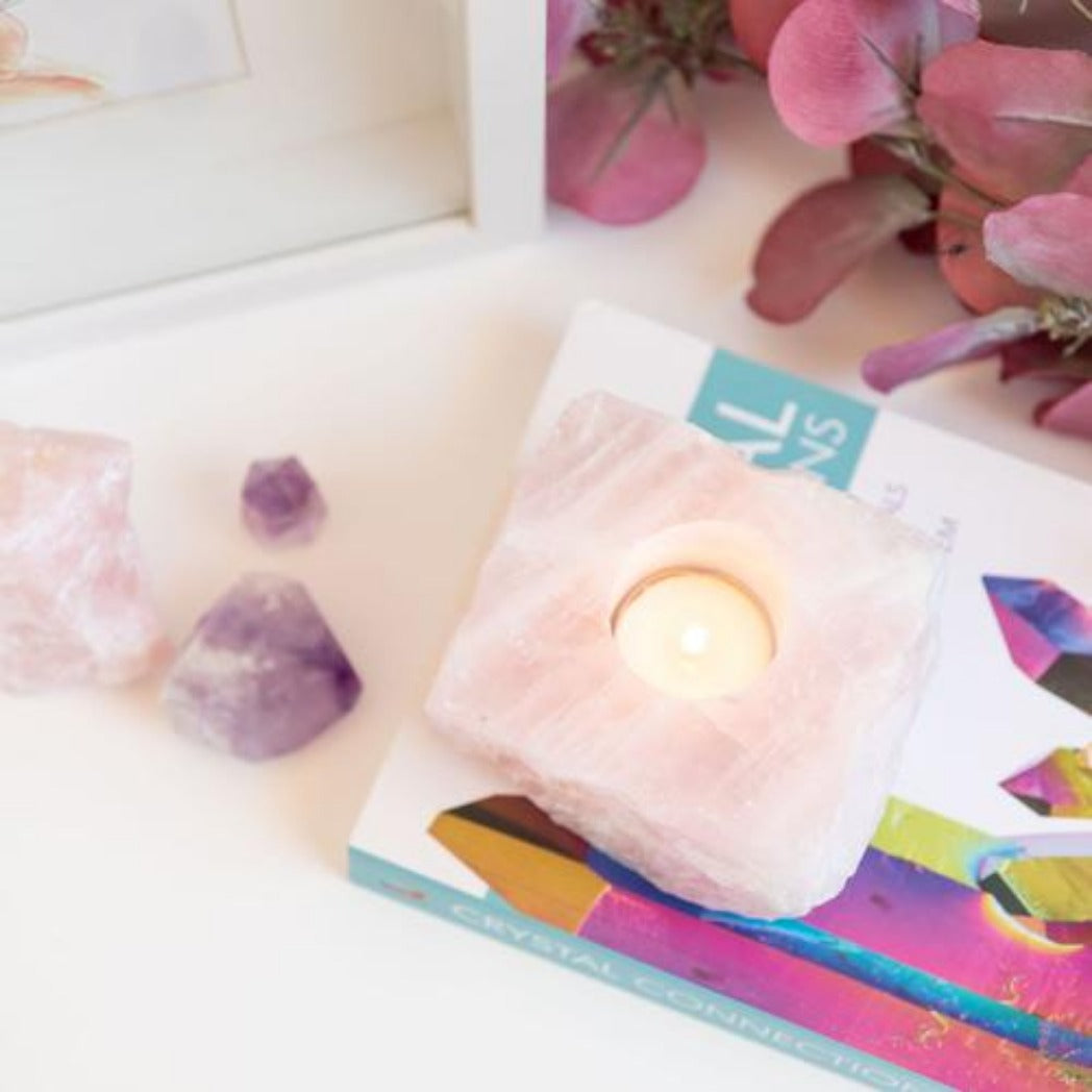 Candle Crystal Homewares Love Rose Quartz Rough Cut. Pale Pink Square cut rose quartz crystal for tealight candle. Crystal Homewares. Candle sits on top of colourful book with amethyst crystal and flowers in background
